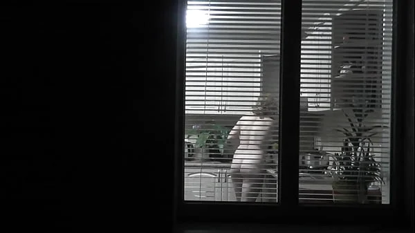 Peeping. Voyeur. Neighbor pervert voyeur in evening on street looks out window as naked sexy neighbor in kitchen prepares dinner for her husband. Naked in public. Naked at home. Family.  Outdoor
